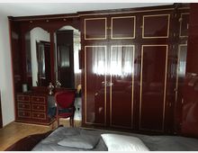 Ein Schlafzimmer mit einem mahagonifarbenen Kleiderschrank und goldenen Zierleisten, einem integrierten Spiegel, einer Schubladeneinheit und einem roten Polsterstuhl. Im Vordergrund steht ein Bett mit grauer Bettwäsche.