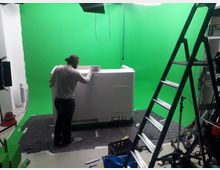 Ein Raum mit einem grünen Hintergrund (Greenscreen), einer Leiter auf der rechten Seite und verschiedenen Ausstattungen wie einer weißen Box in der Mitte, an der eine Person arbeitet. Im Vordergrund liegen Werkzeuge und Kabel, zudem sind Beleuchtungsgeräte sichtbar.