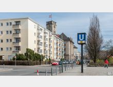 Straßenszene mit mehrstöckigen Wohngebäuden, einem historischen Uhrturm mit Fahnenmast und einem „U4“-Schild einer U-Bahn-Station im Vordergrund. Rechts des Gehwegs stehen kahle Bäume, während Autos entlang der Straße geparkt sind.