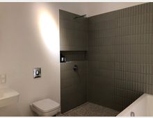 Helles Badezimmer mit weißer Wand, beigefarbenen Fliesen an der Duschwand, einer eingebauten Ablage in der Dusche, einem wandmontierten WC, einem kleinen Waschbecken und einer Badewanne rechts. Der Boden besteht aus Terrazzo.