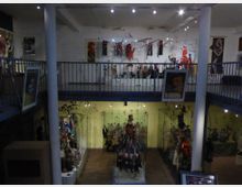 Ein zweistöckiges Puppenmuseum mit Glasvitrinen, in denen verschiedene Marionetten und Puppen ausgestellt sind. Der Raum hat eine umlaufende Galerie im oberen Stockwerk, helle Wände und ist mit Kunstwerken sowie Puppen hängen und stehend dekoriert.