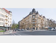 Straßenszene mit einem Eckgebäude im historischen Stil, das beige verputzt ist und von einer großen Kuppel sowie verzierten Türmen gekrönt wird. Im Vordergrund verkehren Autos und Fußgänger, links befindet sich ein modernes Gebäude mit Geschäften im Erdgeschoss, darunter eine Starbucks-Filiale.