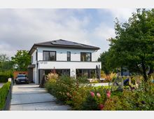 Modernes zweistöckiges Einfamilienhaus mit weißer Fassade, schwarzen Fensterrahmen und einem Schrägdach mit Solarpanels. Vor dem Haus befinden sich eine gepflasterte Einfahrt mit geparktem Auto und ein gepflegter Garten mit Blumen, Sträuchern und einem Kinderspielgerüst.