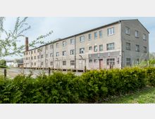 Ehemaliges Industriegebäude mit drei Etagen, grauer Fassade und teilweise beschädigten Fenstern und Türen. Im Vordergrund befindet sich eine grüne Hecke, während im Hintergrund ein Schornstein und andere kleinere Gebäude sichtbar sind.