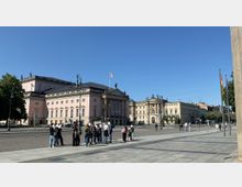 Außenansicht des Bebelplatzes in Berlin mit der Staatsoper Unter den Linden im Vordergrund, einem rosa-farbenen klassizistischen Gebäude mit Säulenfassade. Im Hintergrund sind weitere historische Gebäude zu sehen, während im Vordergrund eine Gruppe von Personen steht, umgeben von einer gepflasterten Fläche und Fahnenmasten.