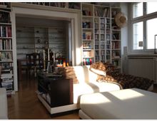 Wohnzimmer mit hellen Möbeln und einem beigeweißen Sofa, daneben ein Bücherregal voller Bücher und CDs. Im Hintergrund ein Essbereich mit Glastüren und Regalen, die mit Geschirr und Gläsern gefüllt sind; rechts ein großes Fenster mit Heizkörper darunter.