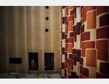 Holzvertäfelte Wand mit mehreren Löchern, darunter zwei rechteckige Öffnungen am unteren Rand und drei runde Löcher in der oberen Hälfte. Rechts ein schwerer Vorhang mit einem orange-roten geometrischen Muster.