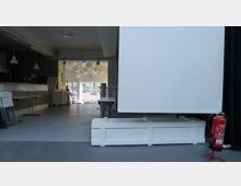 Moderner Innenraum mit einem weißen Podest und einer großen Leinwand im Vordergrund. Im Hintergrund befindet sich ein Raum mit einer Theke, gefalteten Stühlen und hängenden Lampen, sowie ein Ausgang, der auf eine parkähnliche Umgebung führt.