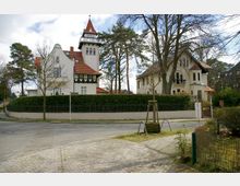 Straßenszene in einem Wohngebiet mit zwei großen historischen Villen im Hintergrund, gekennzeichnet durch weiße Fassaden, rote Ziegeldächer und ein Turmelement an einem der Häuser. Die Gebäude sind von einer Hecke und einem Metallzaun umgeben, während der Vordergrund einen gepflasterten Gehweg zeigt.