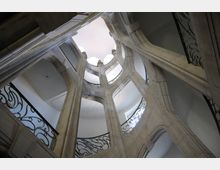 Blick nach oben in einem Treppenhaus mit mehreren Etagen, geprägt von steinernen Rundbögen und schmiedeeisernen Geländern in einem historischen Baustil. Tageslicht dringt durch eine helle Öffnung in der Decke ein.