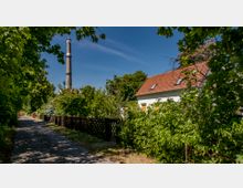 Schmaler Weg mit Bäumen und Sträuchern an beiden Seiten, links ein hoher Schornstein im Hintergrund und rechts ein weißes Haus mit rotem Ziegeldach, teils verdeckt von dichter Vegetation.
