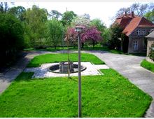 Grüne Hofanlage mit einem zentralen Brunnen auf einer gepflasterten Fläche, umgeben von Rasenflächen, Bäumen und Sträuchern. Im Hintergrund sind historische Gebäude mit roten Ziegeldächern und ein blühender Baum zu sehen.