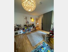 Kinderzimmer mit Holzboden, einem großen grauen Teppich in der Mitte und einer verspielten Deckenleuchte in Federoptik. An den Wänden befinden sich Regale mit Spielzeug und Büchern, eine Spielküche und dekorative Bilder, darunter eine Abbildung einer Giraffe. Ein geöffnetes Tür führt in einen angrenzenden Raum.