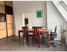 Modern loft-style dining/meeting area with a wooden table, two red-upholstered chairs, and a black office chair on gray flooring. The room has white walls, a slanted ceiling with skylight windows, a cactus in a pot, and an open doorway leading to another room, with large wood wall panels on the left.