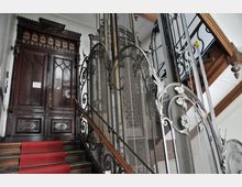 Treppe mit rotem Teppich, die zu einer schweren Holztür mit gotischen Verzierungen führt, flankiert von einem alten Aufzug mit kunstvoll verziertem Metallgitter und Holzgeländer. Der Aufzug ist Teil eines historischen Treppenhauses mit Jugendstil-Elementen.