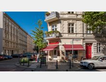 Straßenszene in einer urbanen Umgebung mit einem historischen Eckgebäude und einem kleinen Café mit roten Markisen. Im Vordergrund stehen Tische und Stühle im Außenbereich des Cafés, umgeben von parkenden Autos und Bäumen entlang der gepflasterten Straße.