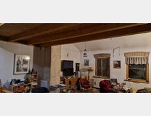 Wohnzimmer in einem rustikalen Stil mit sichtbaren Holzbalken an der Decke, einer weißen verputzten Wand und drei Fenstern mit Holzrahmen und Ziegelbögen. Im Raum befinden sich Möbel, wie Bücherregale, ein Fernseher, ein Tisch und ein Sessel, sowie unterschiedliche Gegenstände, die auf den Oberflächen verteilt sind.