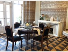 Elegantes Speisezimmer mit rundem Holztisch und vier gepolsterten Stühlen, gedeckt mit Gläsern und Flaschen. Im Hintergrund stehen hohe Fenstertüren, eine gemusterte Tapete und ein weißer Marmorkamin; der Boden ist mit schwarz-beigen Rautenfliesen ausgelegt.