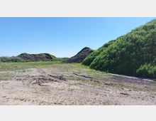 Eine offene Freifläche mit vereinzeltem Grasbewuchs und Spuren im Boden, im Hintergrund zwei grasbewachsene Erdhügel. Die Umgebung wirkt ländlich bei klarem, blauem Himmel.