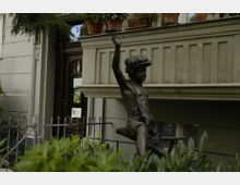 Bronzeskulptur eines Jungen mit erhobenem Arm vor einem historischen Gebäude mit Balkon. Im Vordergrund sind Pflanzen und ein schmiedeeiserner Zaun zu sehen, während im Hintergrund eine Holztür und Fensterelemente des Gebäudes erkennbar sind.