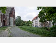 Außenbereich mit einer historischen Kirche aus Back- und Naturstein auf der linken Seite sowie einem beige verputzten Gebäude mit rotem Dach auf der rechten Seite. Dazwischen Grünflächen, Bäume und ein gepflasterter Weg, der parallel zu einer Reihe geparkter Autos verläuft.