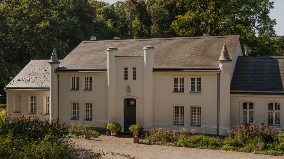 Helles, zweigeschossiges Landhaus mit steilem Schieferdach, symmetrischer Fassade und zentralem Eingang, flankiert von zwei zylindrischen Türmchen und kleinen Eckspitzen. Davor liegt eine Kiesauffahrt mit niedrigen Beeten und Sträuchern, im Hintergrund dichter Baumbestand.