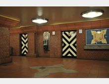 Innenraum eines Gebäudes mit roten Backsteinwänden und einem rotbraunen Fliesenboden. An der Wand befinden sich zwei Türen mit schwarz-weiß gestreiftem Muster, ein öffentliches Telefon und ein Wandplan mit einer Skizze eines Gebäudes. Deckenleuchten sorgen für eine warme Beleuchtung.