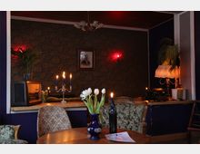 Gemütlich eingerichteter Raum mit floral gemusterten Polstermöbeln, einer mit Tapete bedeckten Wand und gedämpfter Beleuchtung. Auf einem Tisch stehen ein Kerzenständer mit brennenden Kerzen und eine Vase mit weißen Tulpen, daneben eine Tischlampe, eine alte Schreibmaschine und ein Radio als Dekoration.