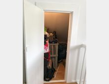 Eine kleine Abstellkammer mit einer offenen weißen Tür, gefüllt mit gestapelten Kisten, Taschen und verschiedenen Gegenständen. Der Raum ist spärlich beleuchtet, und der Boden besteht aus hellem Laminat.