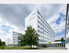 Modernes Gebäudeensemble eines Bildungscampus mit weißer Fassade und großen Fensterfronten; im Vordergrund ein Baum und eine Grünfläche, im Hintergrund ein bewölkter Himmel mit Auflockerungen.