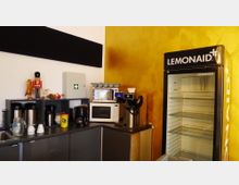 Kleine Büroküche mit grauen Schränken, Kaffeemaschinen, Mikrowelle und einem weißen Kühlschrank mit der Aufschrift "Lemonaid+" vor einer gelben Wand. Im Regal stehen dekorative Figuren sowie ein Erste-Hilfe-Kasten.