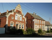 Zwei rote Backsteinhäuser mit detaillierten Fassadenornamenten, eines mit geschwungenem Giebel und Dekorationen. Die Gebäude stehen an einer ruhigen Straße mit Laternen und umgebenden Grünflächen unter klarem Himmel.