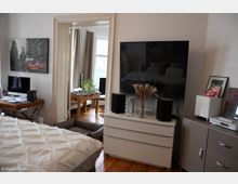 Ein gemütliches Wohnzimmer mit einem großen Flachbildfernseher auf einer weißen Kommode, flankiert von Lautsprechern. Im Hintergrund ist ein angrenzender Raum sichtbar mit einem Tischkicker und großen Fenstern, die viel Tageslicht hereinlassen.