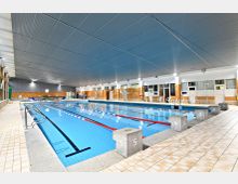 Hallenbad mit einem rechteckigen Schwimmbecken, das mit Bahnmarkierungen und Startblöcken ausgestattet ist. Die Wände sind mit orangefarbenen Kacheln verkleidet, die Decke besteht aus grauen Lamellen, und an der Seite befinden sich große Fenster sowie eine Sitz- und Umkleidebereich.
