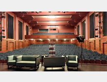 Großer Theatersaal mit mehreren Reihen blauer Sitzplätze, einer Galerie im hinteren Bereich und einer Bühne im Vordergrund. Die Bühne enthält eine Sitzgruppe mit drei Sesseln und einem Tisch, seitlich ist ein Bühnenstrahler zu sehen.