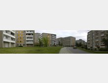 Plattenbau-Wohnsiedlung mit mehreren mehrstöckigen Wohngebäuden aus Beton, teilweise mit gelben und weißen Fassadenelementen. Im Vordergrund eine Wiese und ein Fußweg, rechts eine schmale Straße mit parkenden Autos, umgeben von vereinzelten Bäumen.