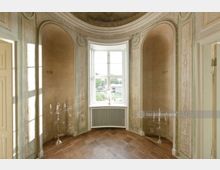 Halbrunder Innenraum mit verzierten Wänden in dezenten Grüntönen, bogenförmigen Nischen und einer großen, weißen Fensterfront, die Tageslicht hereinlässt. Der Boden besteht aus Holzparkett, und silberne Kerzenständer stehen symmetrisch vor den Nischen.