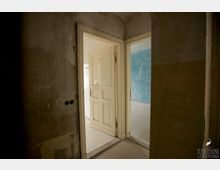 Blick in einen schlichten, renovierungsbedürftigen Innenraum mit abgenutzten, teils beschädigten Wänden. Zwei Türen führen in angrenzende Räume, einer mit weißen Fliesen und der andere mit blauer Wandfarbe.