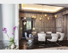 Ein eleganter Essbereich mit einer runden Glasplatte und gepolsterten Stühlen vor einer Wand mit holzartiger Struktur. Im Vordergrund eine Vase mit violetten Orchideen und im Hintergrund ein Ausblick durch ein Fenster sowie eine moderne Hängelampe aus goldfarbenen Kugeln.