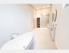 Modernes Badezimmer mit freistehender Badewanne im Vordergrund, einer offenen Dusche mit Glaswand, einer Doppelmarmorwaschbeckenanlage mit runden Spiegeln sowie einem Heizkörper an der Wand; im Hintergrund ist ein kleiner Saunabereich zu sehen.