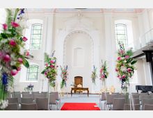 Heller Innenraum eines historischen Gebäudes mit hohen weißen Wänden, großen Bogenfenstern und einer verzierten Nische im Hintergrund. Der Raum ist für eine Hochzeit oder Zeremonie geschmückt, mit Stuhlreihen, einem roten Läufer in der Mitte und Blumenarrangements in Rosa, Weiß und Grün auf hohen Ständern.