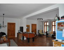 Wohnbereich mit Holzboden, weiß gestrichenen Wänden und mehreren Holztüren. Der Raum ist mit großen Holzrahmenfenstern, einem Tischkicker, einem Sofa, einem Sessel und einem Hängeschrank ausgestattet.