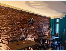 Gemütlicher Innenraum einer Gaststätte mit einer rustikalen roten Backsteinwand, schwacher Beleuchtung und schwarzem Holzmobiliar. Im Vordergrund stehen mehrere Tische mit Stühlen, während die Wand von einem dekorativen Schild und Lichtakzenten geprägt ist.