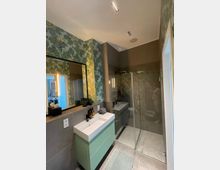 Modernes Badezimmer mit einer gläsernen Dusche rechts, einem grünen Waschtischunterschrank mit weißem Waschbecken links und einer großen Wandspiegelfläche. Die Wände sind teilweise mit gemusterter Tapete im Pflanzen- und Zitronendesign versehen, der Boden besteht aus neutralen Fliesen.