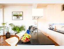 Moderne Küche mit heller Einrichtung, einer schwarzen Arbeitsplatte und integrierter Kochinsel; an der Wand hängen Küchenutensilien, und Zimmerpflanzen sowie eine Schale mit Obst dekorieren den Raum. Ein gerahmtes Bild und Holzboden ergänzen die schlichte, wohnliche Atmosphäre.