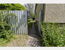 Schmaler Außenweg neben einem Wohnhaus mit heller Klinkerfassade, rechts mit Regenfallrohr und dichtem Unkrautbewuchs. Links steht ein hoher, grauer Wellblechzaun mit Eingang, dahinter Bäume und ein kleiner Gartenbereich.