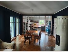 Wohnraum mit offener Küche, Holzboden im Fischgrätmuster und dunklen Wänden. Links große Fenster mit Vorhängen, ein hölzerner Esstisch mit Stühlen in der Mitte, im Vordergrund rechts ein weißer, runder Kachelofen und im Hintergrund eine helle Küche mit modernen Elementen und direktem Blick ins Grüne.