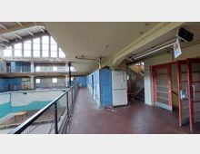 Ein verlassenes Hallenbad mit leerem Becken, blauer und weißer Fliesenverkleidung sowie hohen, bogenförmigen Fenstern. Im Vordergrund befindet sich ein Flur mit blauen Umkleidekabinen, einer Treppe und abgesperrtem Bereich.