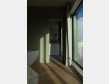 Ein modernes Zimmer mit hellgrünen Wänden, einem Holzboden und großem Fenster, das natürliches Licht hereinlässt. Im Hintergrund ist eine offene Tür zu einem Badbereich mit einer weißen Badezimmerkommode und Fliesenboden sichtbar.