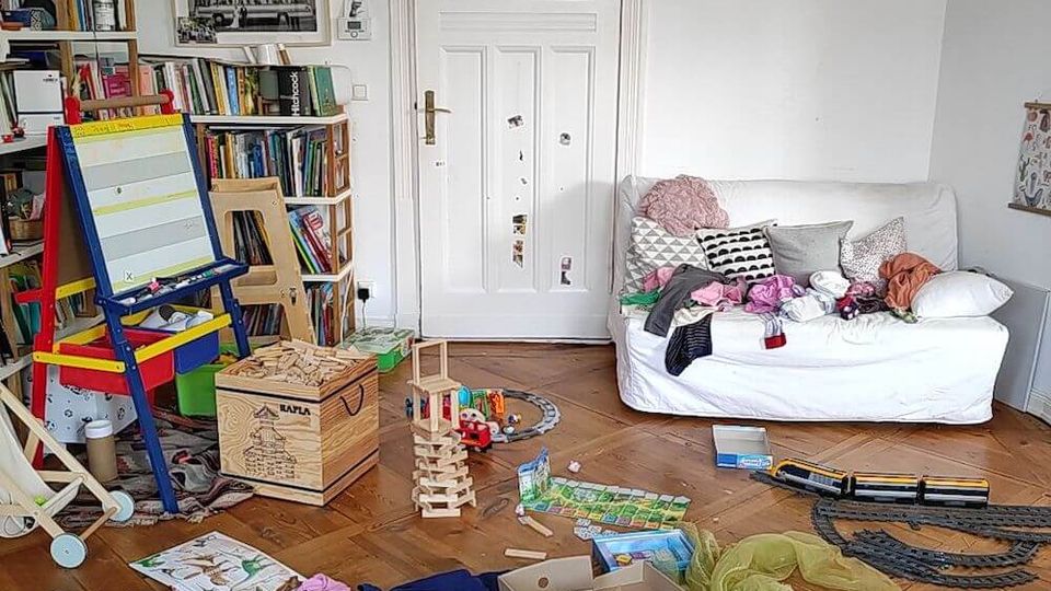 A cluttered living room with a white wooden door, a couch with scattered cushions, and shelving filled with books. The wooden floor is covered with toys, books, and game pieces, suggesting an active play area.